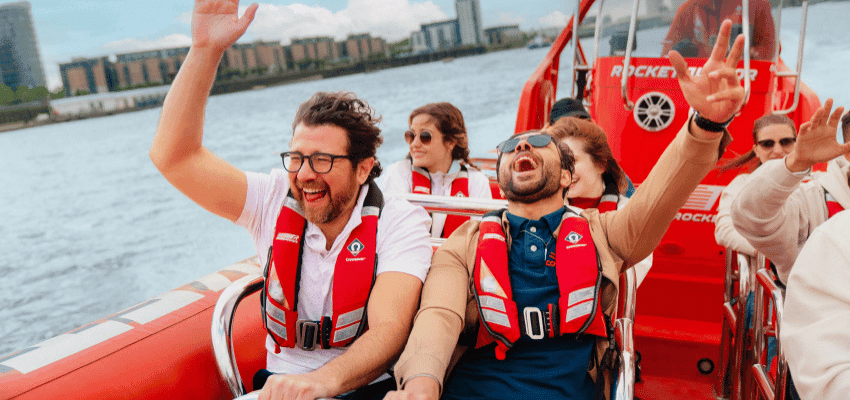A group of people wearing life jackets are joyfully riding a speedboat on a river. Two men in the front are laughing and raising their arms in excitement. The background shows waterfront buildings under a partly cloudy sky.