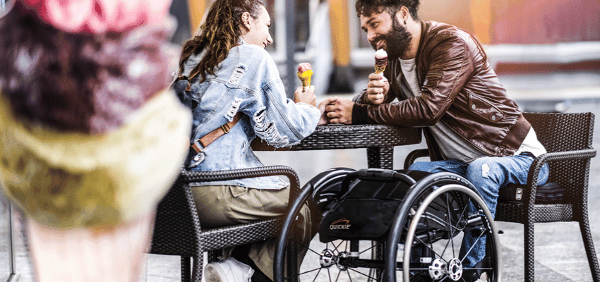 Young man using a manual wheelchair while enjoying ice creams with a partner outdoors, representing accessible days out, inclusive living and mobility discounts for disabled people in the UK.