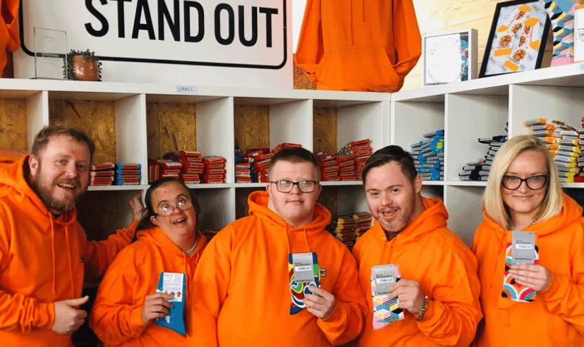 Five people wearing matching orange hoodies stand in front of shelves filled with colourful socks. They are holding up pairs of socks and smiling at the camera. Above them, a sign reads "STAND OUT." The background includes various display items and decorations.