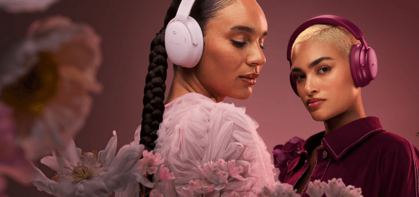 A dramatic close-up of two people wearing Bose over-ear headphones in a floral setting with pink and purple tones. One person wears light pink/lilac headphones with their hair in a braid and a sheer pink ruffled top. The other wears deep magenta/plum headphones and a dark purple collared garment, looking directly at the camera.