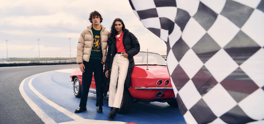 Two models in Tommy Hilfiger clothing stand beside a red car on a race track with a large chequered flag in the foreground. Image used to promote Purpl discounts and accessible fashion choices for people with disabilities and long term health conditions.