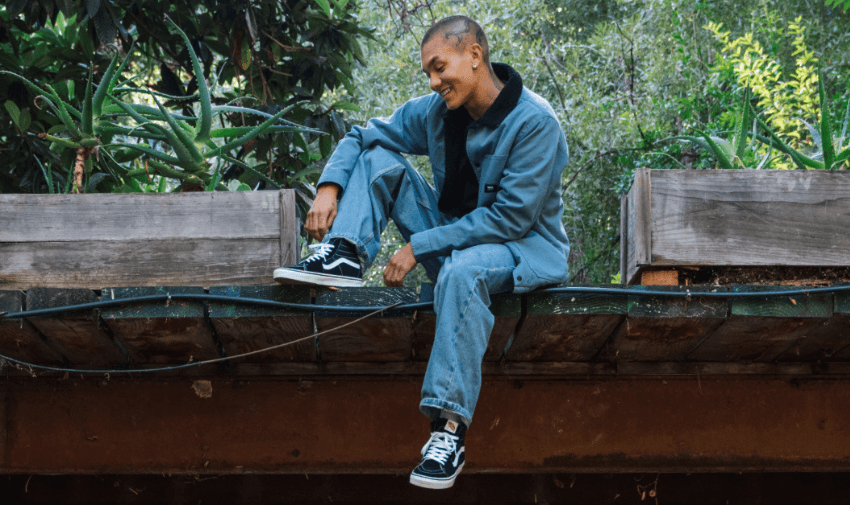 Smiling person sitting on a raised wooden platform outdoors, wearing a blue outfit with classic black and white Vans trainers, highlighting inclusive fashion choices and disabled discount offers on casual footwear in the UK.