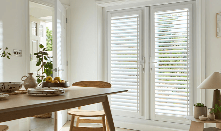 Bright kitchen-dining space with white plantation shutters on French doors, letting in natural light. Perfect for stylish light control and privacy. Ideal for accessible home improvements and energy-efficient window treatments for disabled people.