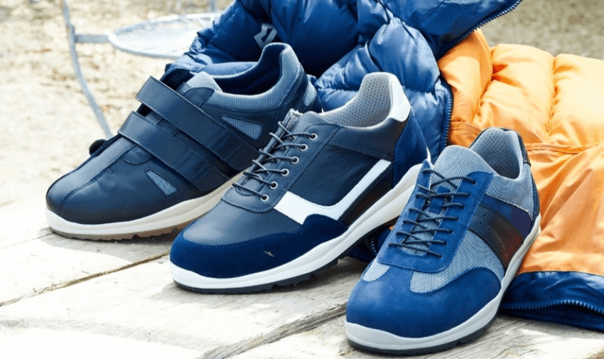 Three pairs of wider fit men’s trainers in shades of navy and blue placed beside padded outdoor jackets, featuring options with velcro straps, classic laces, and breathable materials for accessible, supportive footwear.