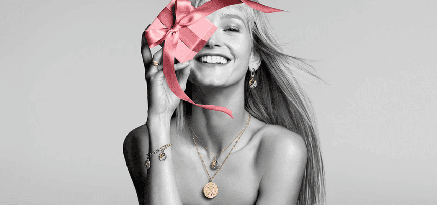 A woman smiling while holding a pink ribbon-tied gift box near her face, wearing layered gold jewellery, supporting the Pandora offer with Purpl discounts for people with disabilities and long term health conditions.