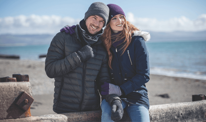 Smiling couple in winter knitwear by the sea, Heat Holders Purpl discounts for disabilities and long term health conditions, mobility discounts and offers.