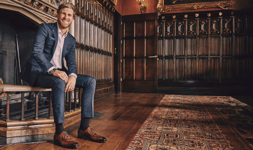 A man in a suit and smart brown shoes sits on a bench near a fireplace in a room with dark wooden paneling and an ornate rug.