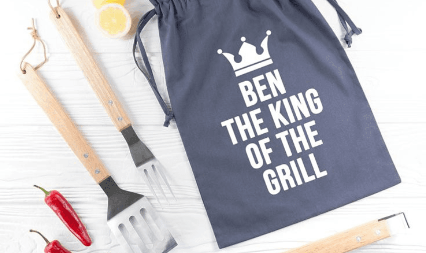 Personalised navy-blue BBQ tool bag with ‘Ben The King Of The Grill’ printed in white next to three wooden-handled stainless steel barbecue tools, red chillies, and lemon slices, showing inclusive gift ideas and fun Father’s Day presents for disabled people in the UK.