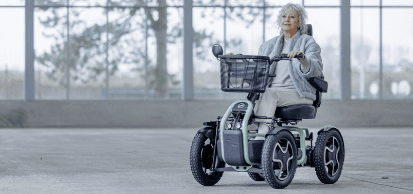 Older woman with mobility needs riding an all-terrain mobility scooter indoors, promoting accessible mobility solutions and UK mobility discounts for people with long term health conditions.