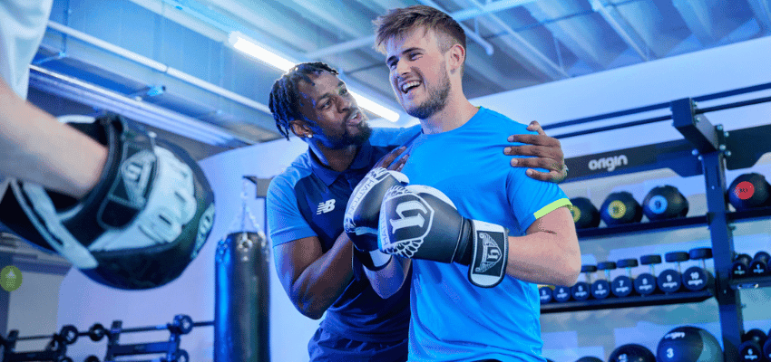 Boxing trainer coaching a smiling man wearing gloves at The Gym Group gym, representing accessible fitness programmes and shopping discounts for disabled people in the UK