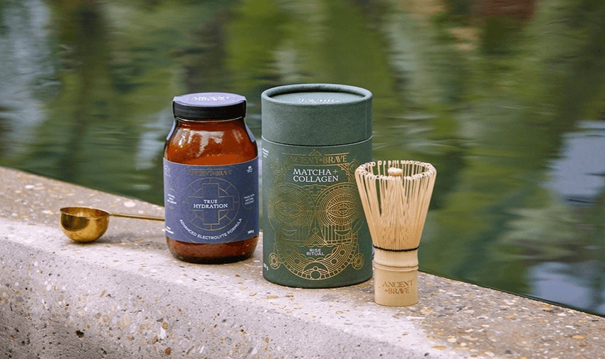 Ancient + Brave wellness bundle beside water, featuring a jar of True Hydration, a canister of Matcha + Collagen, and a bamboo matcha whisk – premium supplements for hydration and skin health