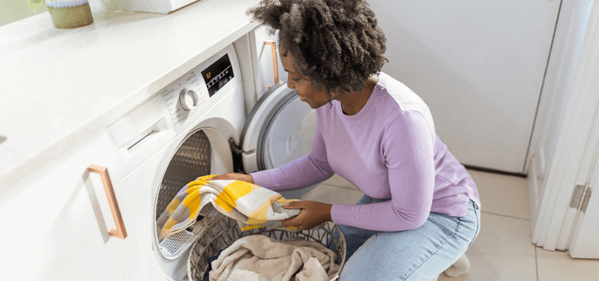 A woman in a lavender sweater is kneeling to load laundry (a yellow and white towel) into a washing machine.
