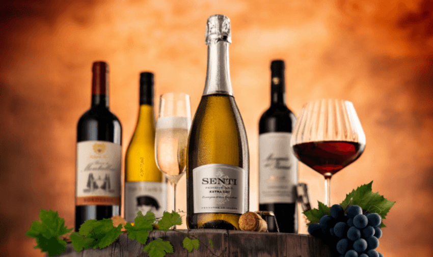 A premium collection of wine bottles and glasses, including red, white, and sparkling wine, arranged on a wooden barrel with grapes and leaves, showcasing affordable indulgence and wine bundle savings for disabled people in the UK.