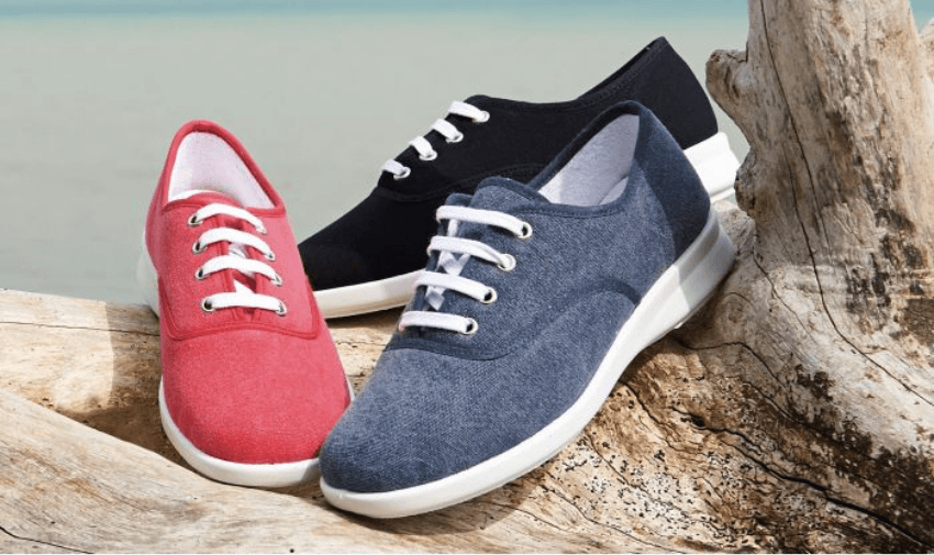 Three pairs of lace-up canvas shoes with white soles displayed on driftwood by the seaside, one red, one denim blue, and one black. Designed for comfort and extra width, suitable for people with mobility needs or foot conditions.