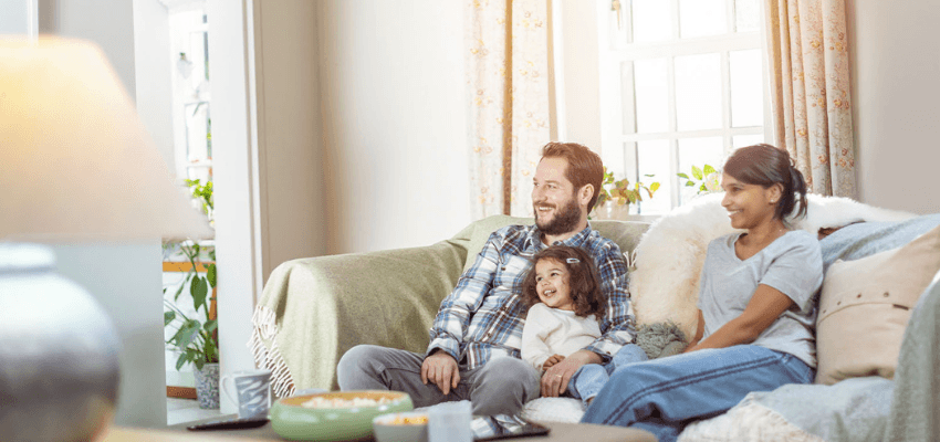 A family of three, including two adults and a young child, smiling while sitting on a sofa in a brightly lit living room with a bowl of popcorn nearby.