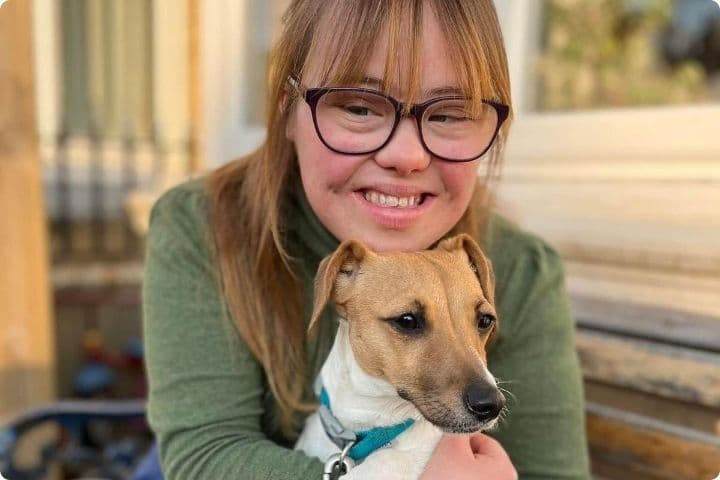 Florence Halliwell is a UK based land artist with Down's syndrome, she has dark blonde long hair with a fringe and wears glasses, she has a sage green long sleeved top on and is smiling and hugging a small terrier dog who has a blue colour on