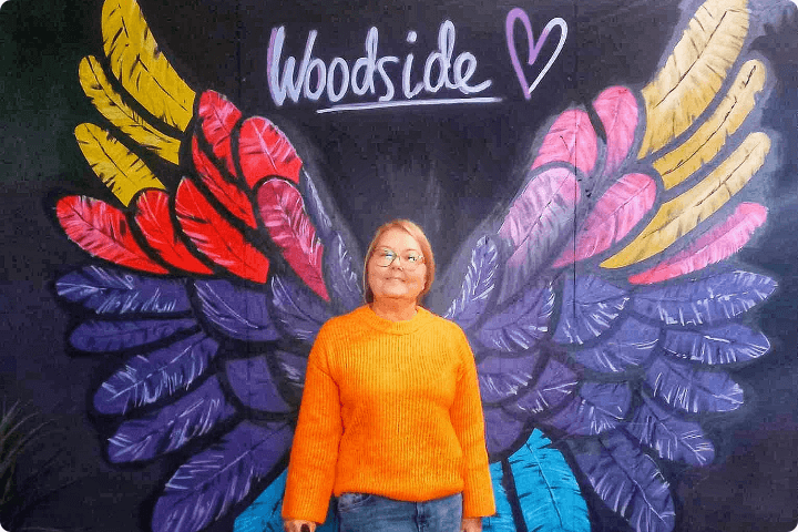A woman in her 30s is wearing an orange jumper and uses a mobility cane, she is standing in front of a painted mural on a wall of colourful wings with the word woodside and a heart, she has blonde hair in a bob and is wearing glasses and smiling