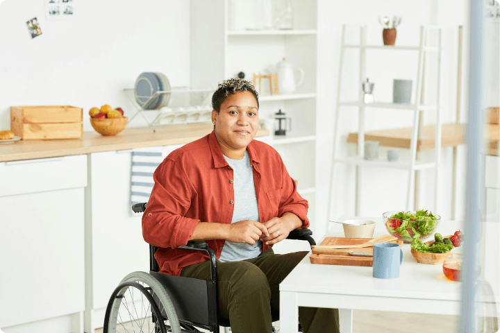 A woman with short cropped hair sits at a kitchen table preparing food, she is a wheelchair user and is wearing a red shirt and white tshirt