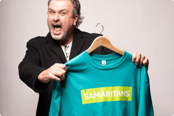 Jon McClure is a white man in his 40s, he has greying hair and a gold front tooth, he is holding up a green samaritans t-shirt and has his mouth open as though he is shouting