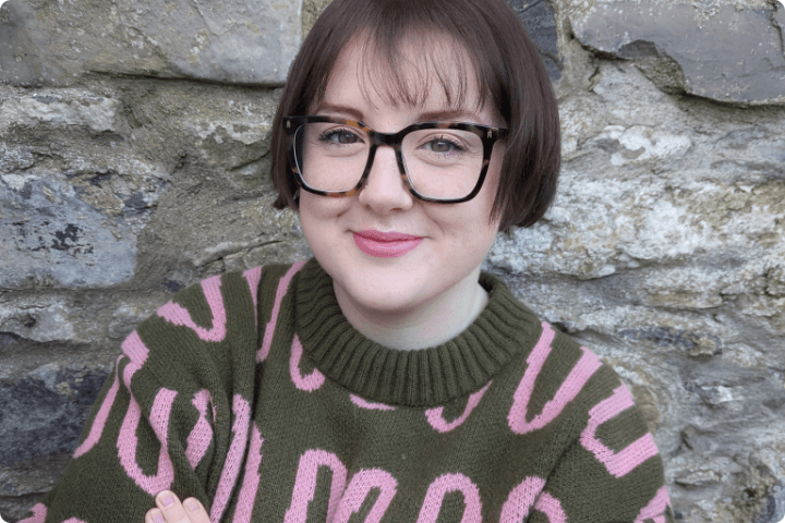 Rebecca is a white woman, she has short brown hair in a bob with a fringe and wears glasses, she is wearing a brown jumper with pink squiggles on it.