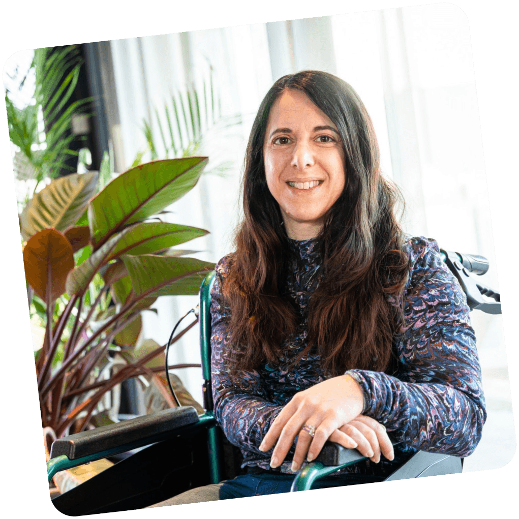 Woman with long dark hair sitting in a wheelchair, smiling at the camera. She is wearing a patterned long-sleeve shirt and sitting in a bright room with green plants in the background.
