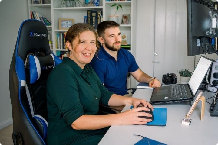 Leah, a woman in her 30s wearing a green shirt dress, and Finn, a man in his twenties in a blue polo shirt, sitting together at a desk. In the background is a large bookcase holding numerous books, dictionaries, pot plants, and an award.