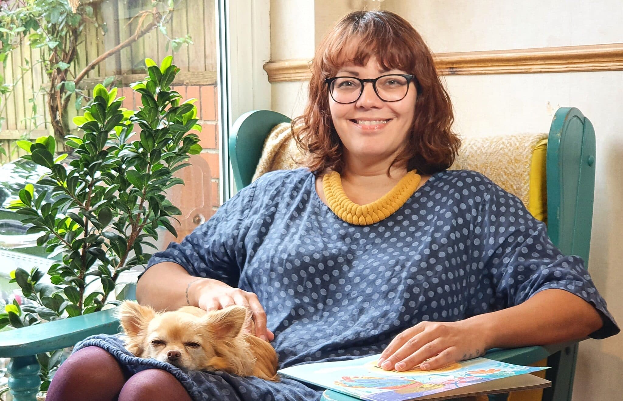 Sam Cleasby is a woman in her early 40s of Indian and British heritage, she has brown curly hair and glasses and she is sat on a chair with a chihuahua on her lap, she is wearing a blue dress and a yellow necklace and there is a plant in the background